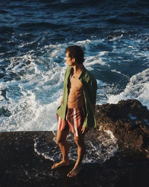Model Louis Baines standing on rocky shore wearing open olive green shirt and striped shorts by the sea for Zara