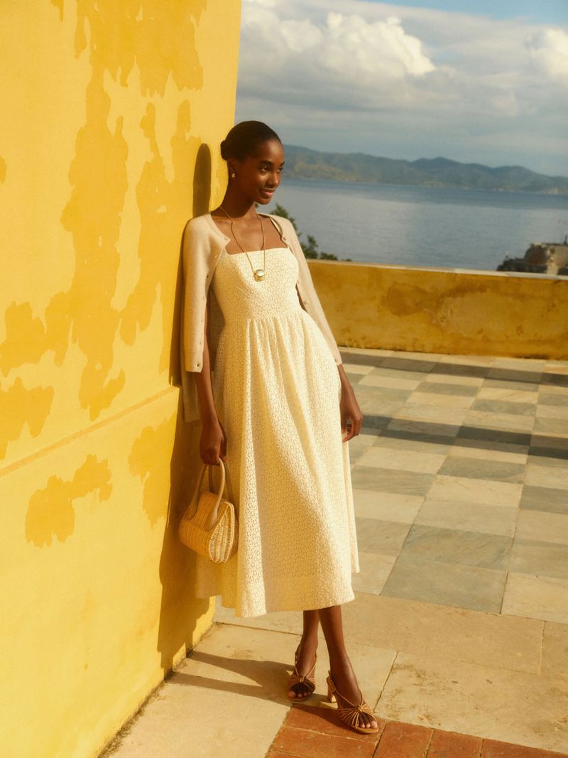 Woman in a white dress poses near a yellow wall and overlooks the ocean. She is holding a handbag, wearing sandals and jewelry.