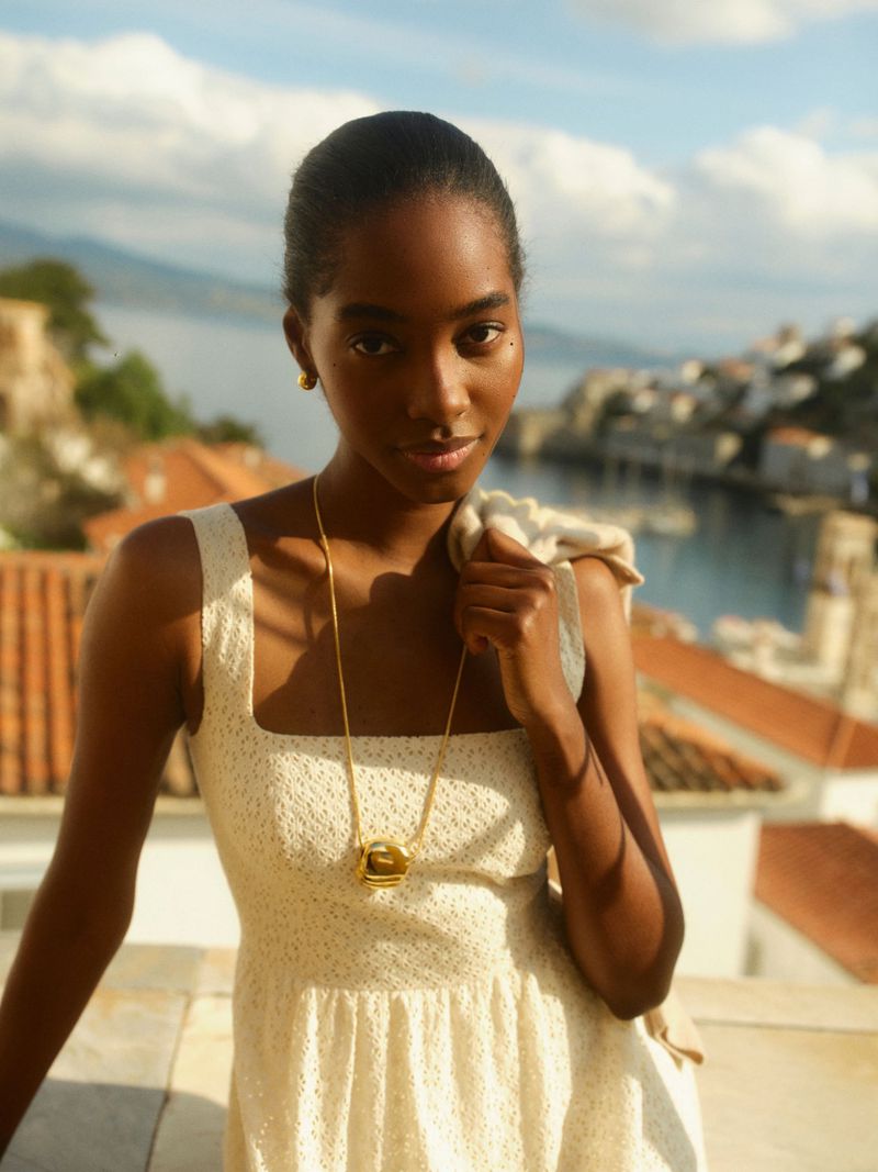 Model in white dress wearing gold necklace against coastal town backdrop