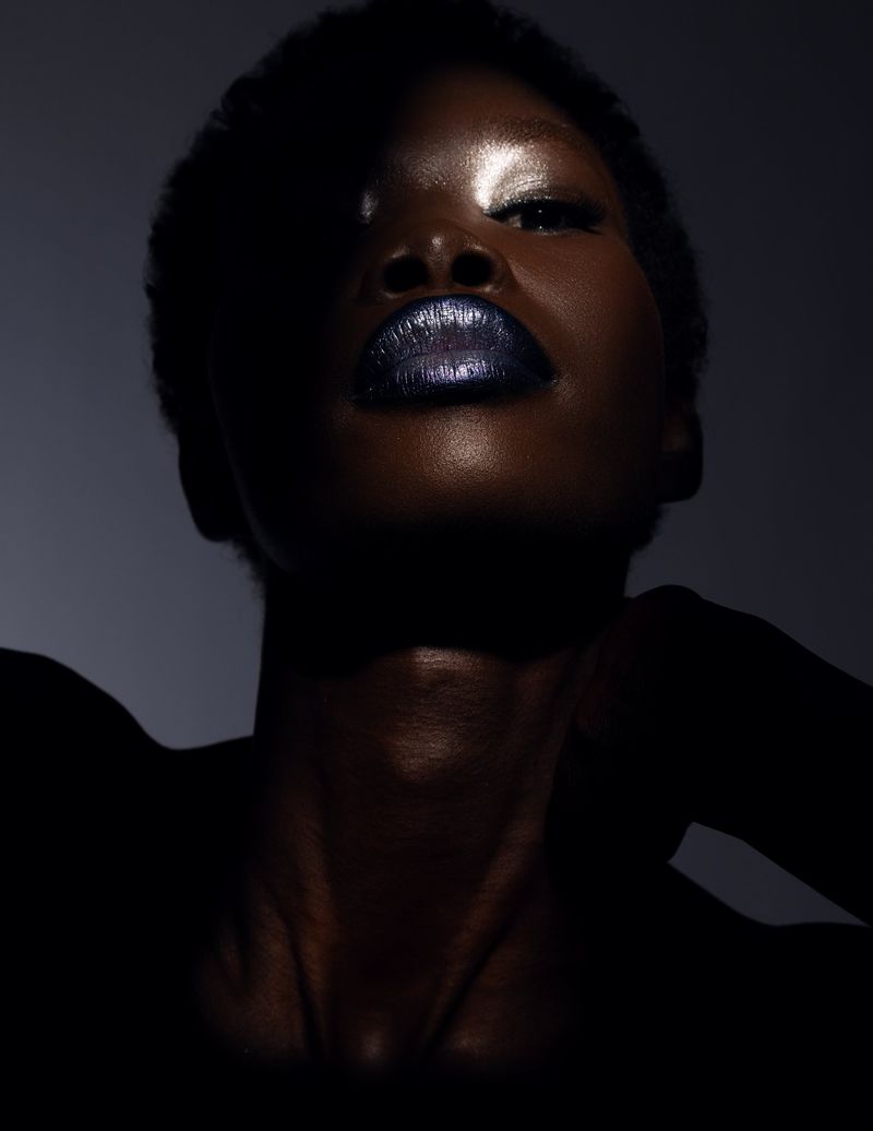 Close-up studio shot of Black adult female with dark, glossy lips, dramatic lighting, and wet skin effect on a dark gray backdrop.