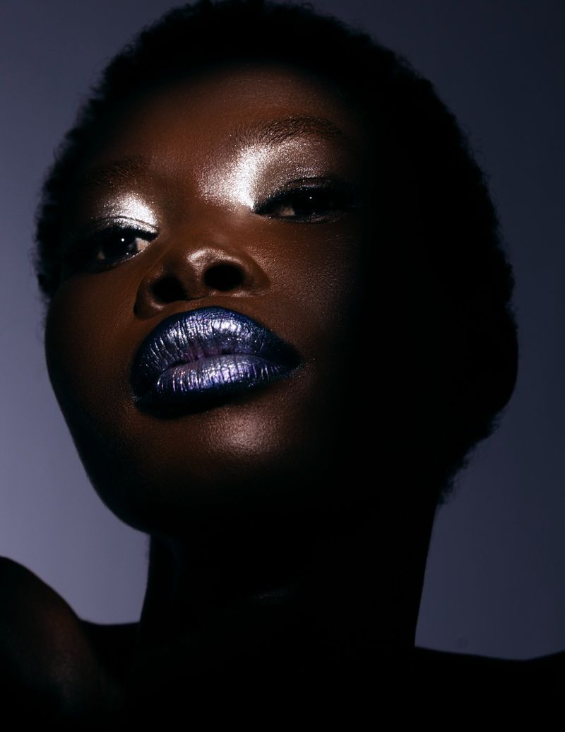 Close-up studio portrait of an adult female model with dark skin and glittering blue lipstick, looking up with bright eyeshadow.