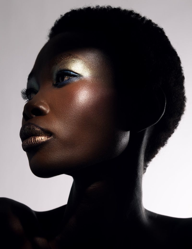 Close-up portrait of a black woman with dark skin, bold gold eye makeup, and matching gold lipstick against a gradient gray background.
