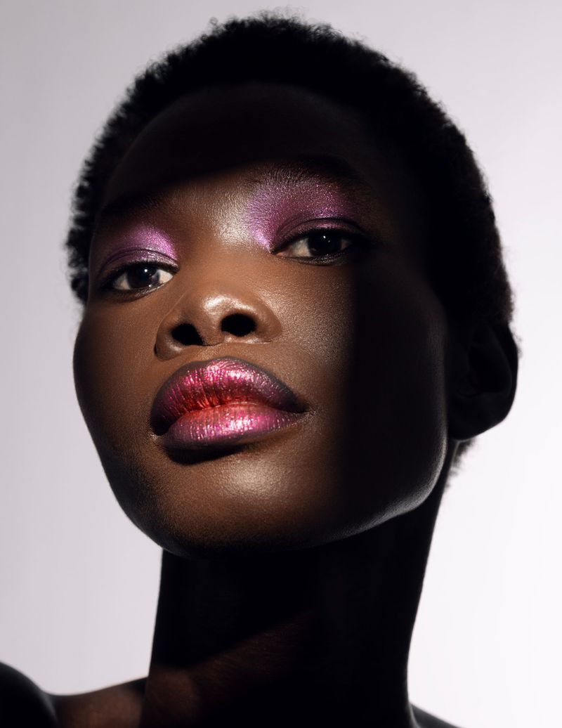 Close-up portrait of a Black woman with vibrant pink and purple eye shadow and glossy lips against a gradient grey background.