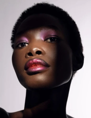 Close-up portrait of a Black woman with vibrant pink and purple eye shadow and glossy lips against a gradient grey background.