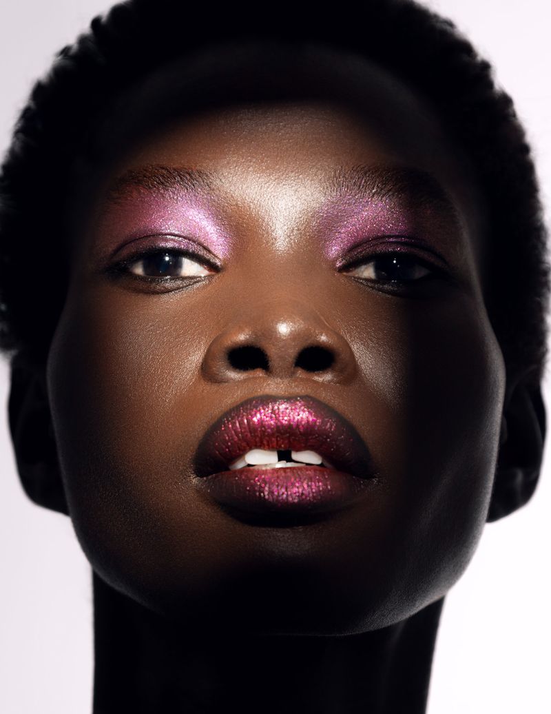 Close-up portrait of a Black woman with pink and purple eyeshadow and glittery pink lips against a stark white background.