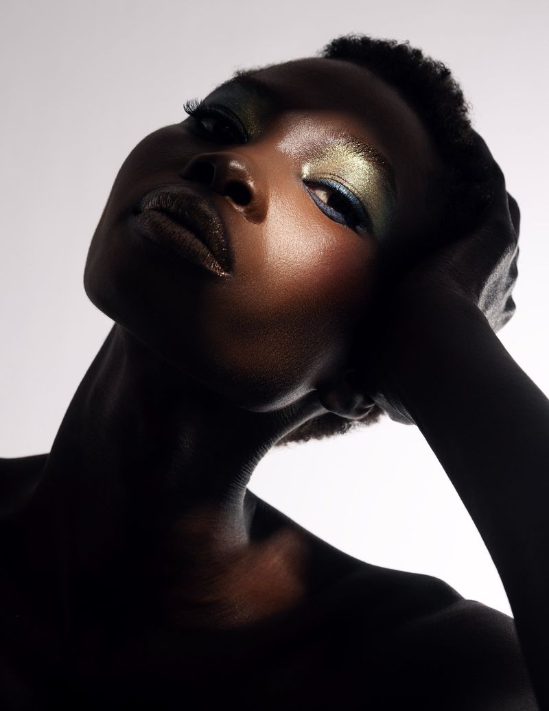 Close-up portrait of a Black woman with shimmering metallic eyeshadow and glossy dark lips against a white background.