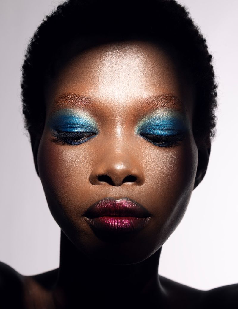 Close-up portrait of a Black woman with vibrant blue eyeshadow and metallic plum lipstick against a white backdrop.