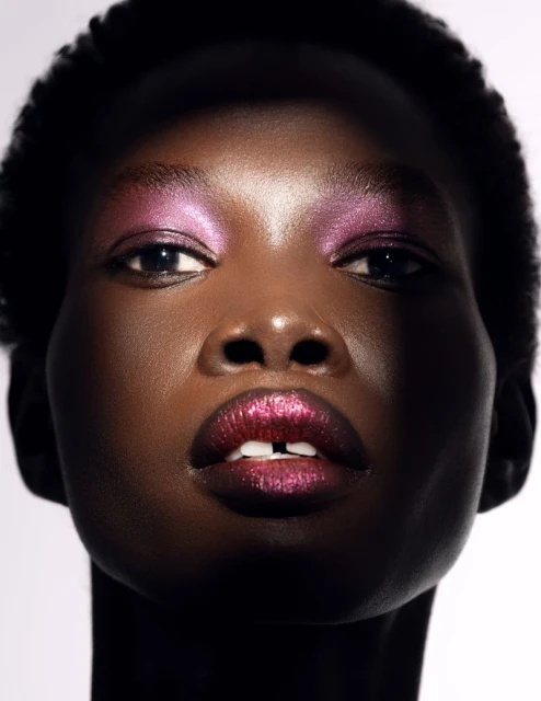 Close-up portrait of a black woman with metallic pink eyeshadow and glossy lipstick against a white background.