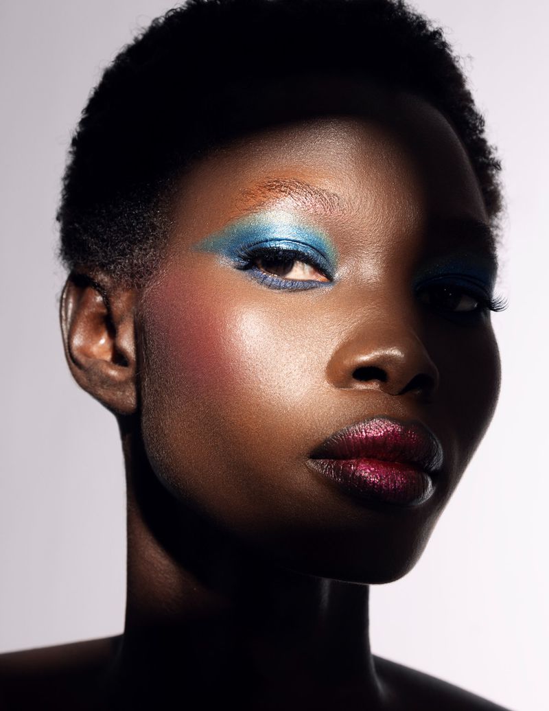 Close-up portrait of a Black model with blue eye shadow and dark lipstick against a plain white background.