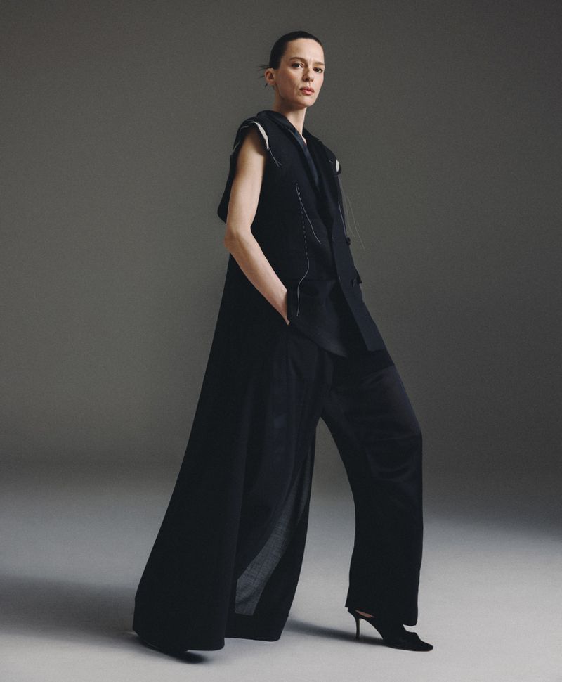 Model wearing a long black vest and wide trousers standing against a seamless grey studio background