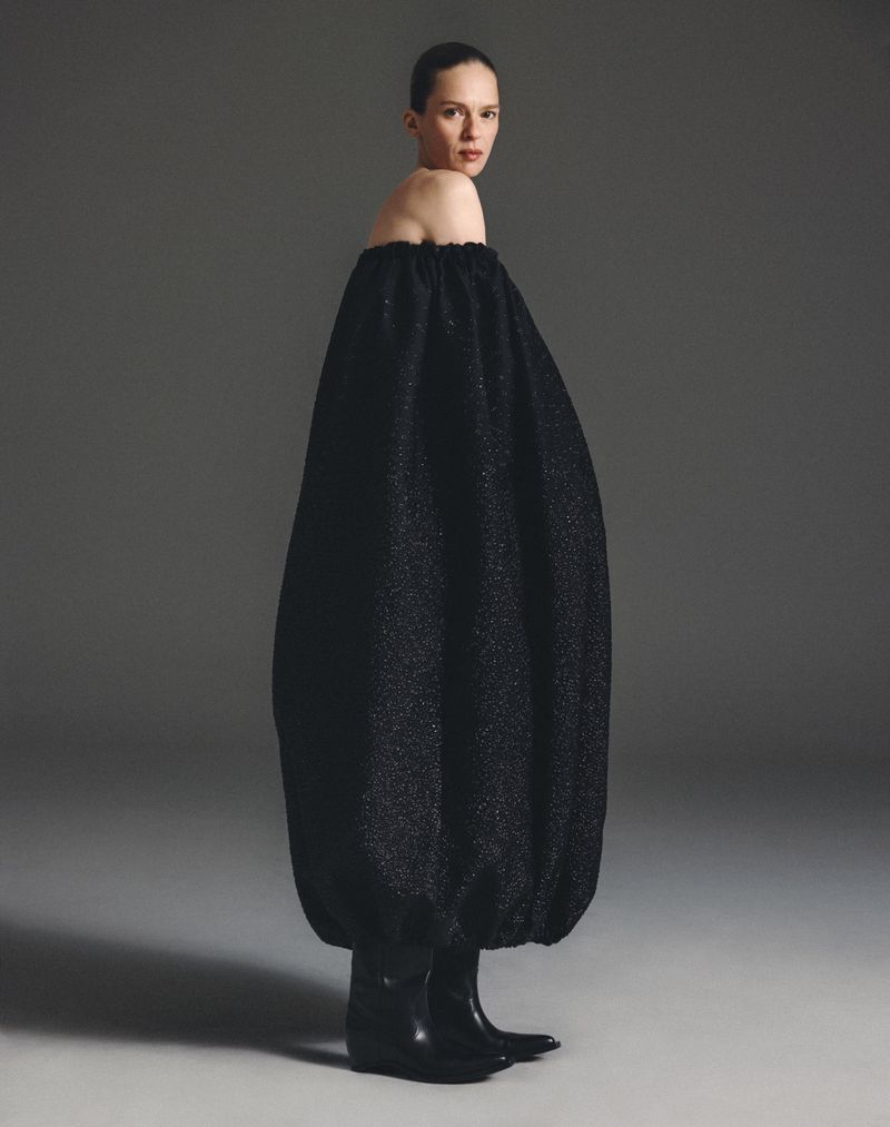A model showcases a black off-the-shoulder dress with a unique bubble silhouette, paired with black heeled boots in a studio setting.