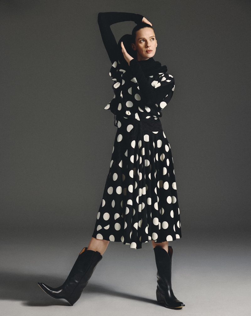 Model Elise Crombez wearing a black and white polka dot dress and black cowboy boots against a seamless grey studio background.