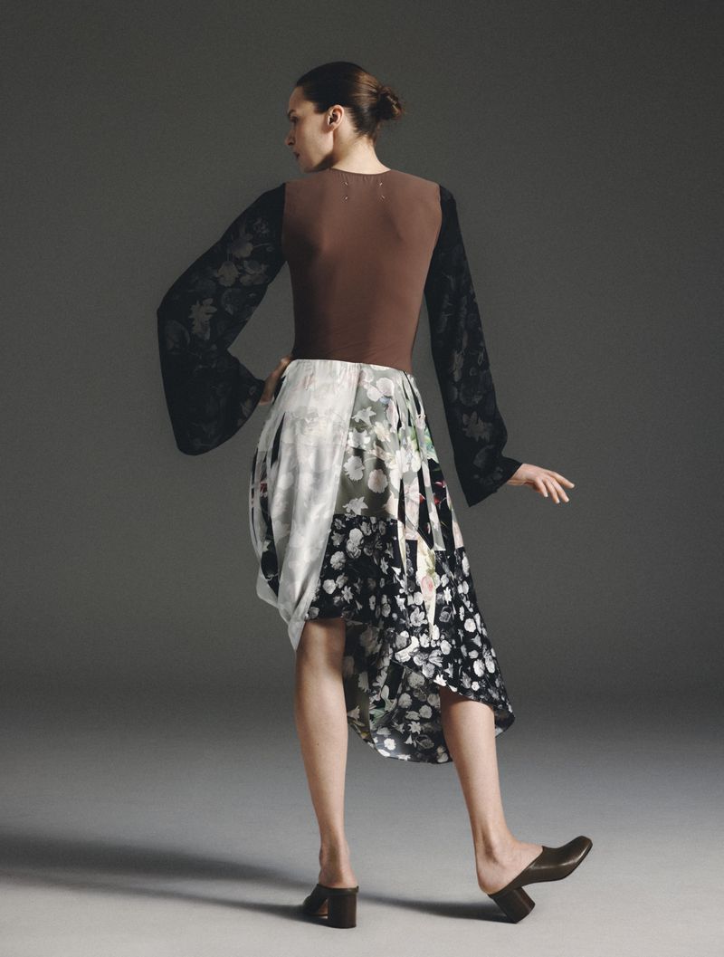 Model wearing brown top with black patterned sleeves and floral skirt viewed from behind against a grey background
