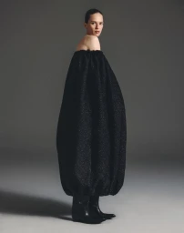 Model Elise Crombez wearing a voluminous black off-shoulder dress and boots against a gradient grey studio backdrop.