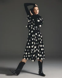 Model Elise Crombez wearing a black and white polka dot dress and black cowboy boots against a seamless grey studio background.