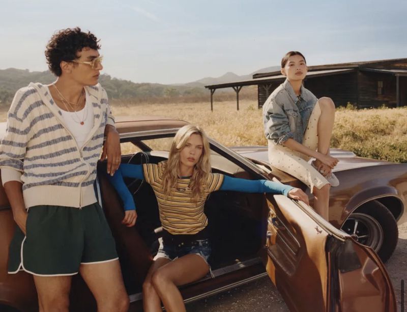 Three fashion models pose on a vintage car in a field against blue sky; one woman sits on the hood, another on the door, and a man stands.