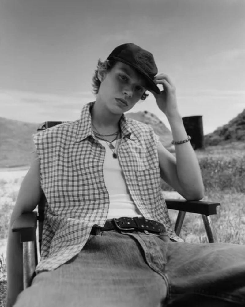 Monochrome portrait of a young male model in a cap, plaid vest, and jeans seated outdoors.