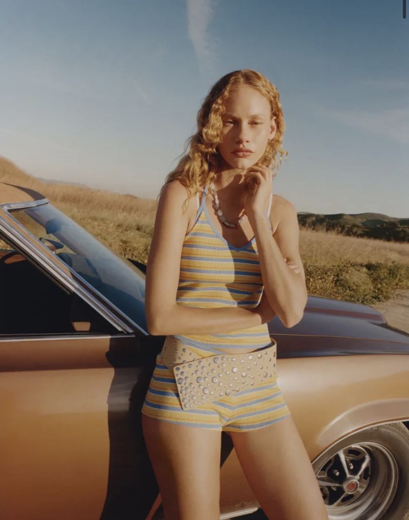 Model with blonde curly hair leaning against a classic car wearing a striped top and studded belt outdoors.