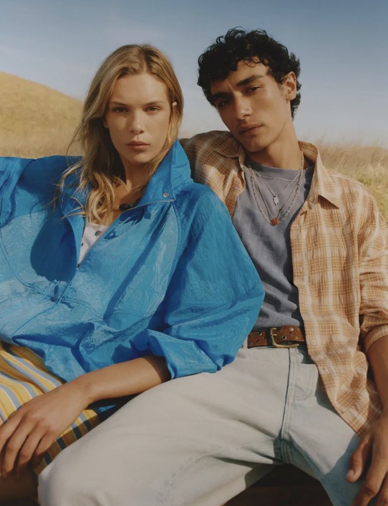 Two models outdoors against a dry grassy hillside, wearing casual blue and plaid apparel for a Zara campaign.