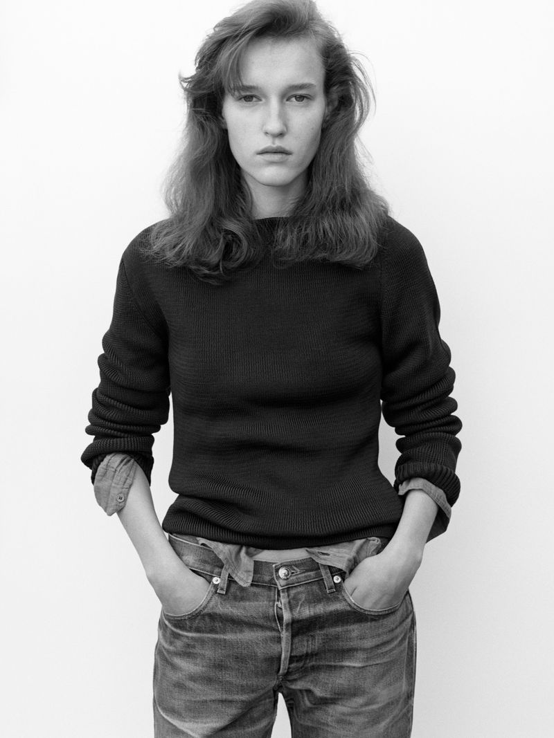 Indoor portrait of a young adult woman with neutral expression wearing a dark sweater and jeans against a white backdrop.