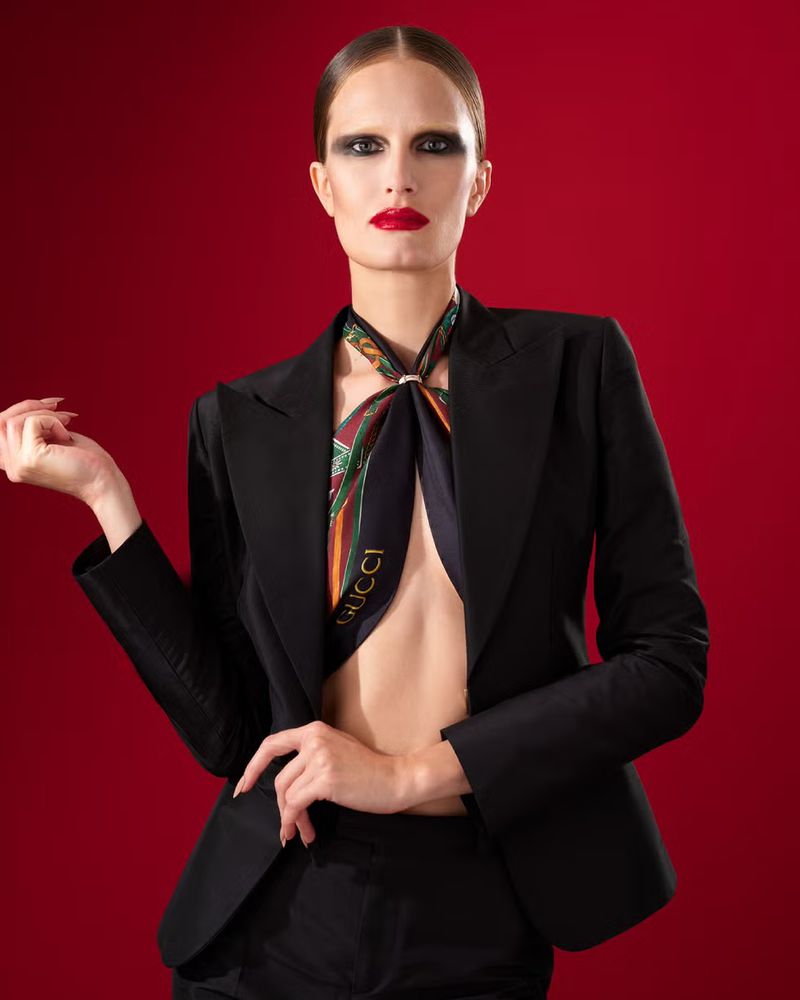Model wearing a black blazer and patterned silk scarf against a rich red background for Gucci.
