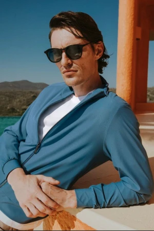 Male model wearing sunglasses and a blue zip-up jacket outdoors against a clear sky and distant hills for SCAPPINO SS 2026.