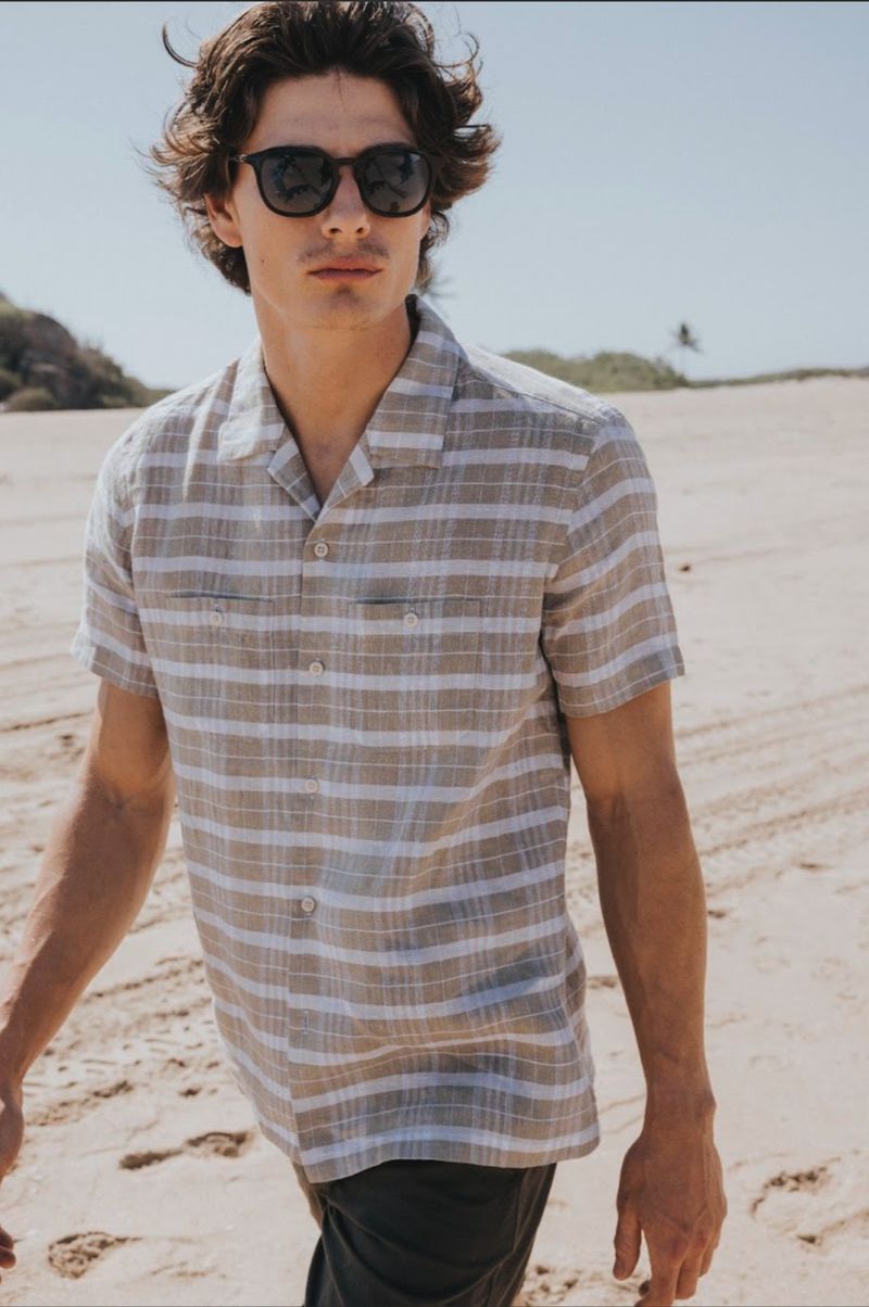 Male model wearing sunglasses and a striped short-sleeve shirt walking on a sandy beach.