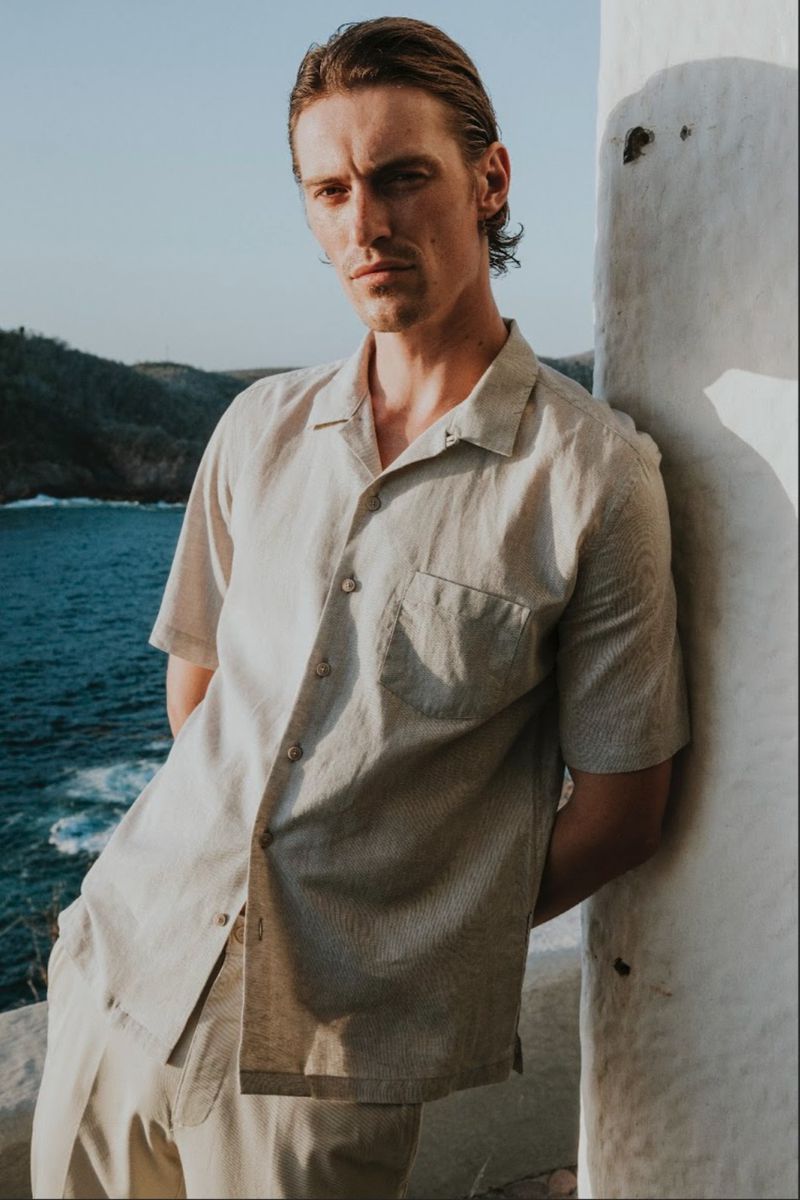 Model Myles Crosby wearing a cream linen shirt leaning against a white pillar by the ocean for Scappino SS 2026