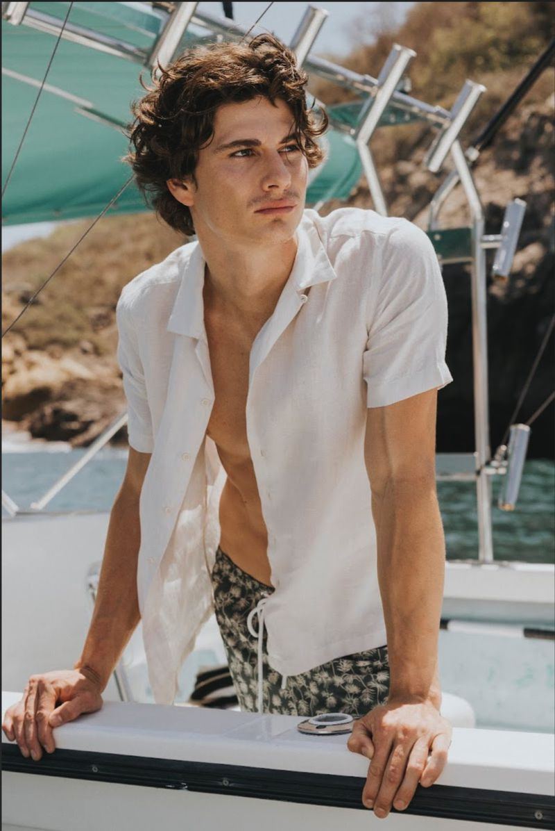A young male model with dark curly hair wearing an unbuttoned white shirt and patterned swim shorts aboard a boat.