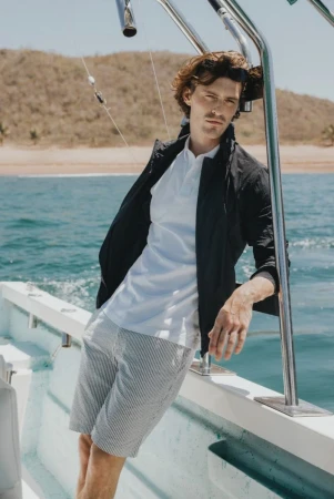 A man leaning against a boat railing wearing a white polo shirt, dark jacket, and patterned shorts on the water.