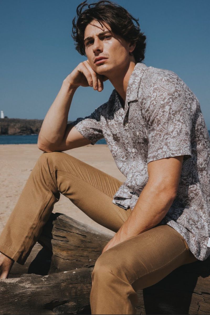 Male model wearing a patterned short-sleeve shirt and khaki trousers seated outdoors near the ocean