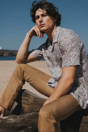 Male model wearing a patterned short-sleeve shirt and khaki trousers seated outdoors near the ocean