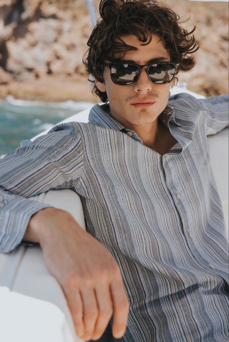 Model wearing dark sunglasses and a striped button-down shirt seated on a boat deck against a coastal backdrop.
