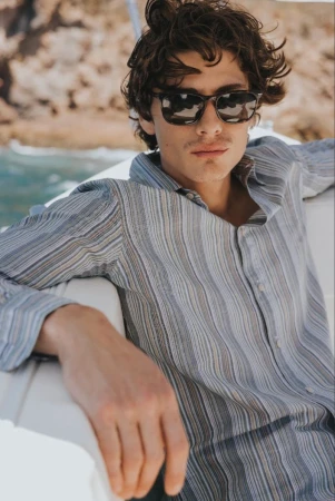 Model wearing dark sunglasses and a striped button-down shirt seated on a boat deck against a coastal backdrop.