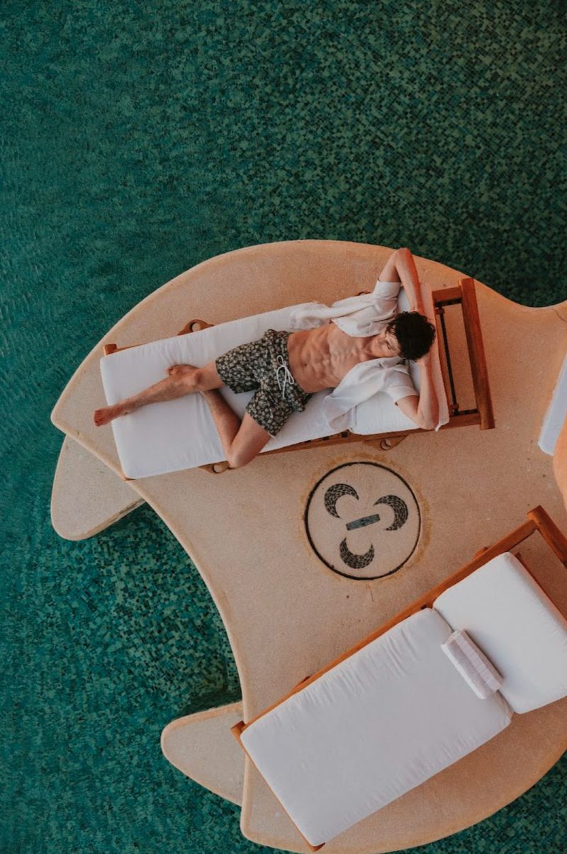 Aerial view of a male model relaxing on a wooden lounger by a turquoise swimming pool for the Scappino SS 2026 campaign.