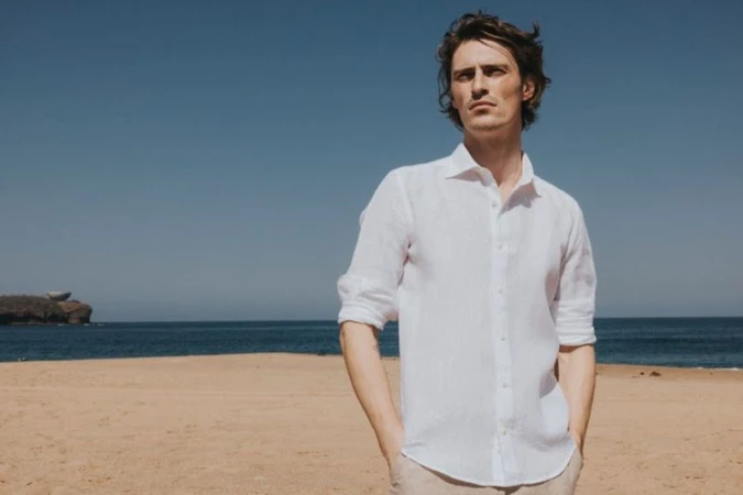 Male model wearing a white linen shirt posing on a sandy beach under a clear blue sky