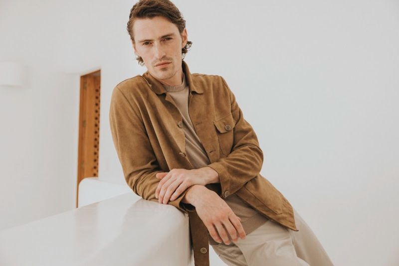 Male model wearing a brown suede jacket leaning against a white structure in a bright minimalist interior.