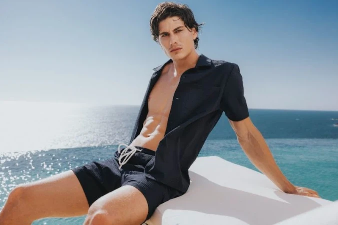 Male model wearing unbuttoned dark shirt and swim trunks seated against an ocean view for SCAPPINO SS 2026 campaign.