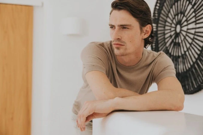 Man wearing a beige t-shirt leaning against a white surface inside a bright room with a round black decorative wall hanging.