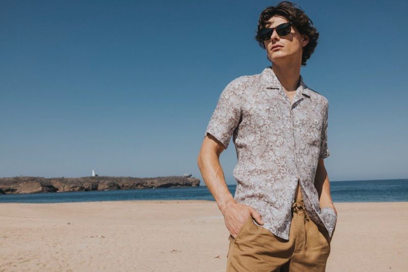 Male model wearing sunglasses and patterned shirt on sandy beach with ocean background