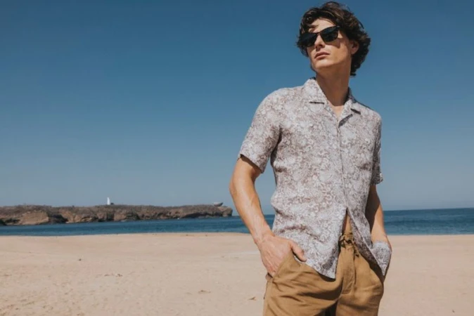 Male model wearing sunglasses and patterned shirt on sandy beach with ocean background