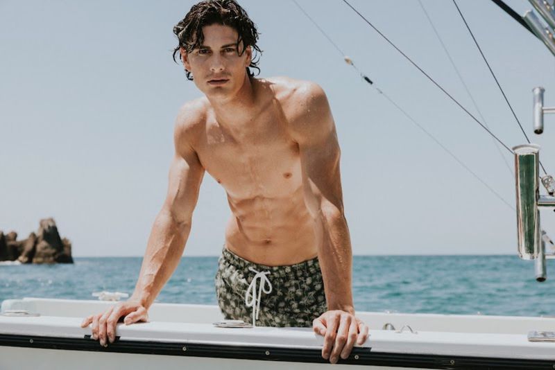 Shirtless male model leaning on the edge of a boat on the ocean wearing patterned swim trunks