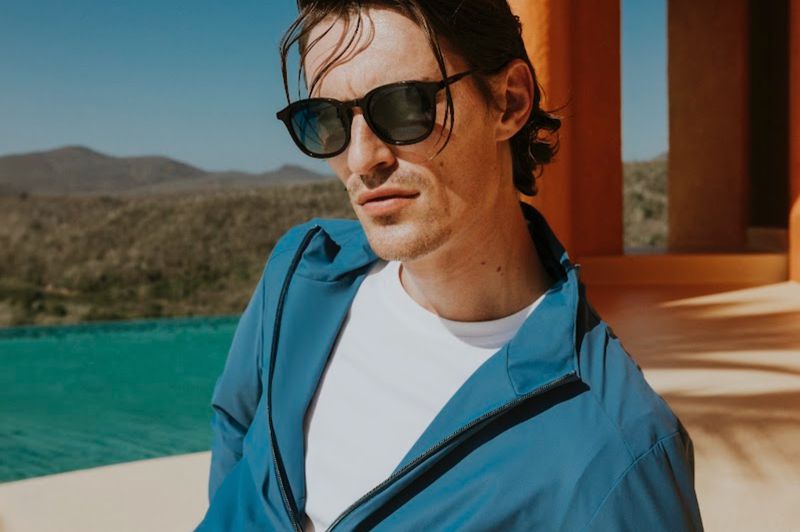 Close-up portrait of a male model wearing dark sunglasses, white t-shirt, and blue zip jacket outdoors near a resort pool.