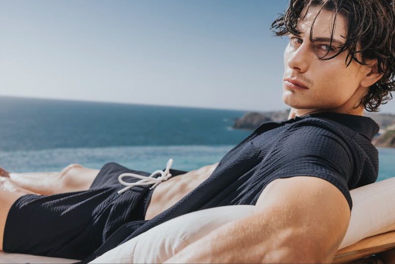 A man wearing dark swim shorts and a matching polo shirt lounging poolside with the ocean in the background for Scappino SS 2026.