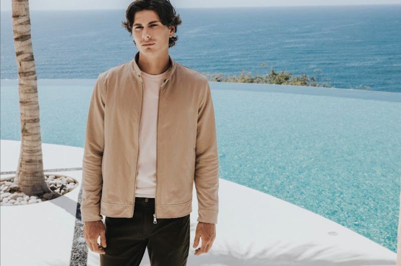 Male model wearing a beige jacket and dark trousers standing by an infinity pool overlooking the ocean