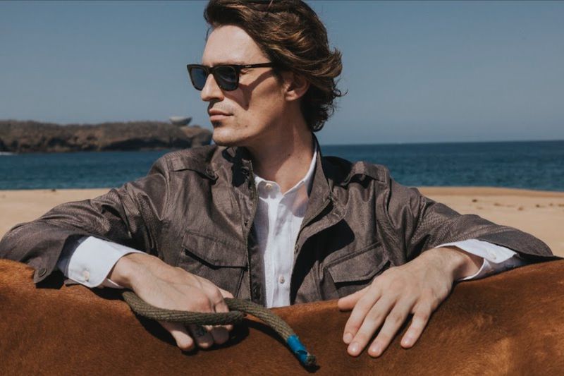 Man wearing sunglasses and a grey jacket outdoors by a sandy beach and ocean for Scappino SS 2026 campaign.