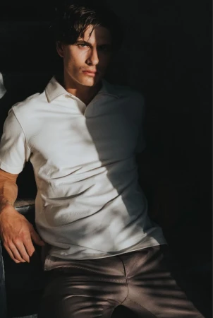 A man wearing a white polo shirt and brown trousers poses in harsh directional light with shadow patterns
