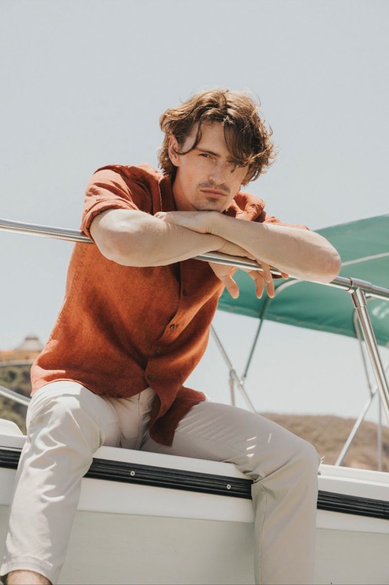 Male model in an orange shirt and white trousers leaning on a boat railing against a clear sky background