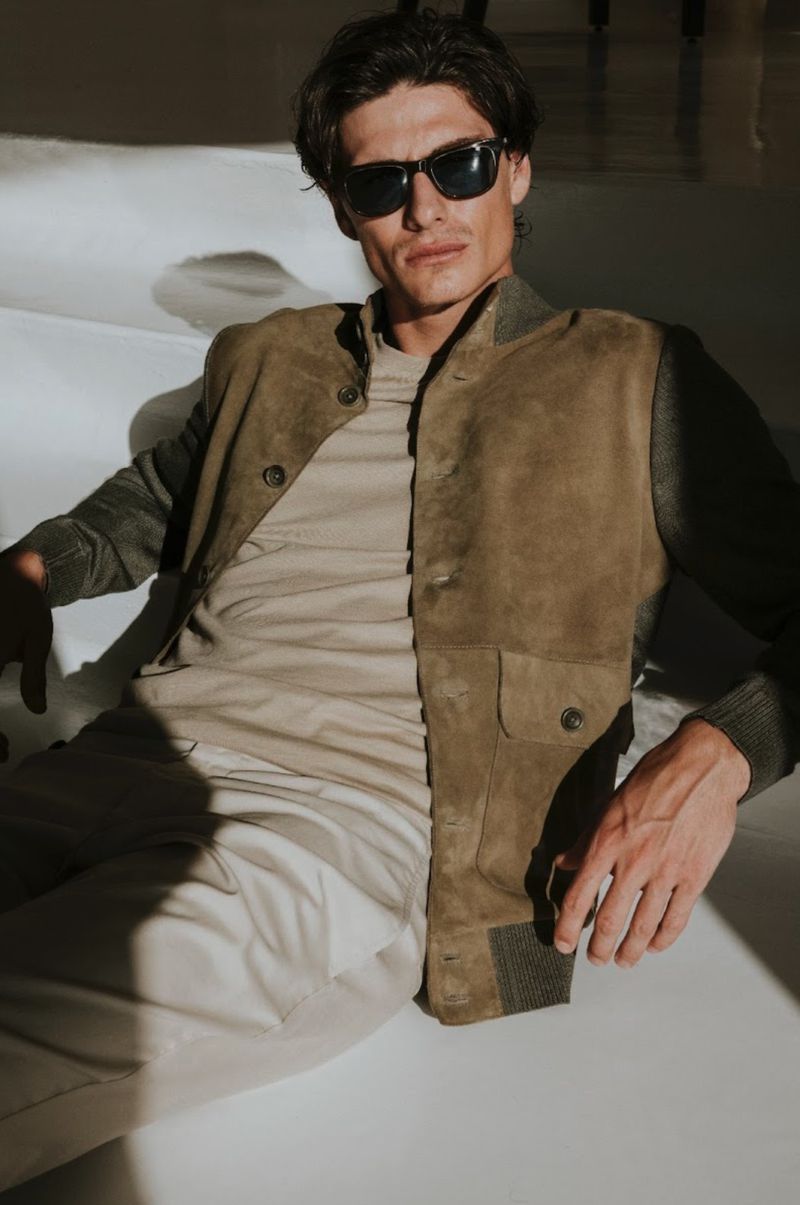 Male model wearing dark sunglasses, an olive suede jacket, beige t-shirt, and light trousers reclining on a white set.