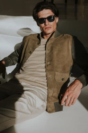Male model wearing dark sunglasses, an olive suede jacket, beige t-shirt, and light trousers reclining on a white set.